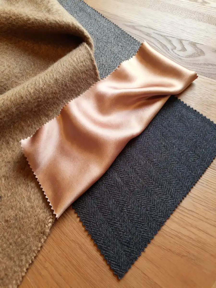 Fabric swatches featuring tan wool, rose gold silk, and charcoal grey herringbone on a light wood surface for textile design.