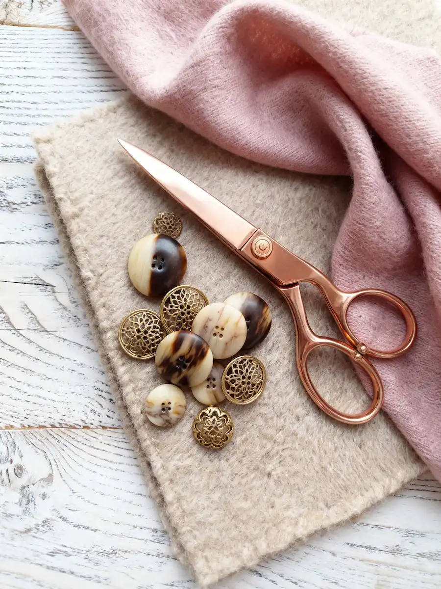 Rose gold sewing scissors resting on beige wool fabric next to decorative gold and tortoiseshell buttons.