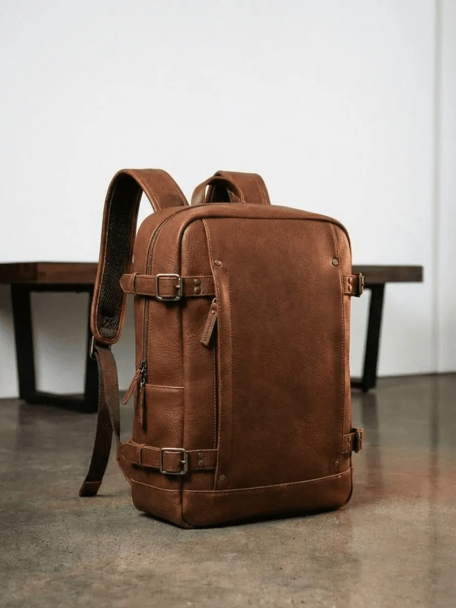 Capra Leather Tamarao backpack in brown full-grain leather with adjustable side buckle straps and a minimalist design, displayed on a concrete floor.2026 luxury gift guide.