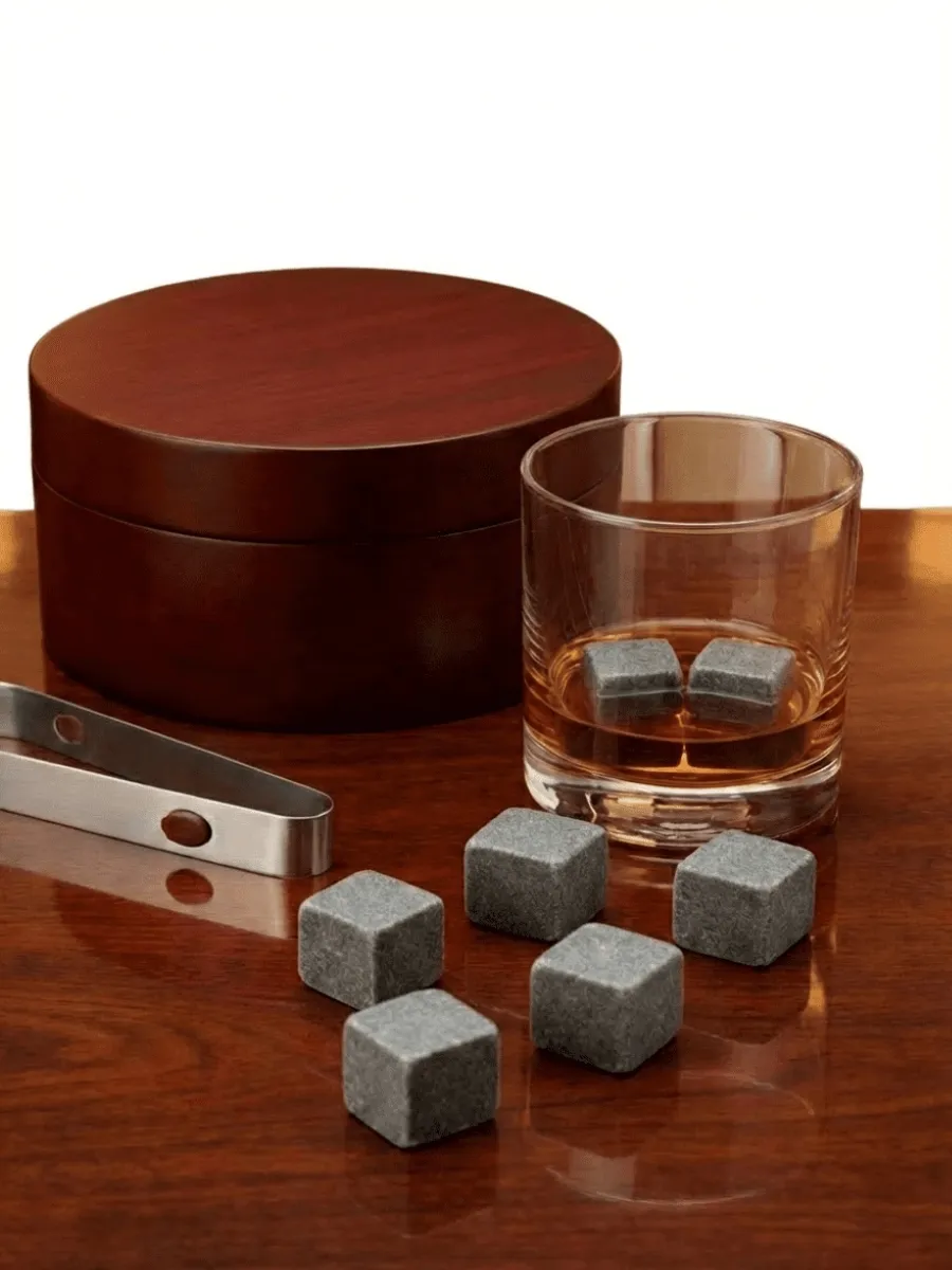 Set of six grey granite whiskey stones on a dark wood table next to a glass of bourbon and a circular wooden storage case. 2026 luxury gift guide.