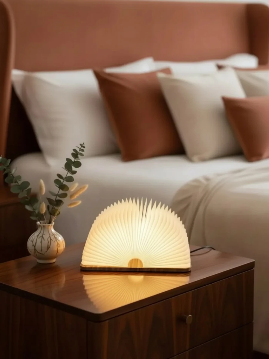 Wooden Book Lamp Review: Affordable Luxury for Your Cozy Home Nook A fanned-open wooden book lamp glowing warm light on a wooden nightstand in a bedroom with terracotta pillows.