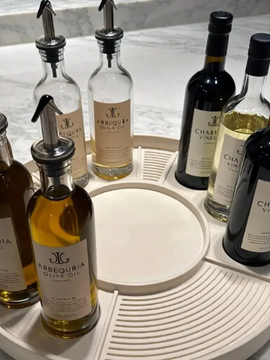 A cream-colored lazy susan organizer holding olive oil and vinegar bottles with minimalist labels on a marble countertop.