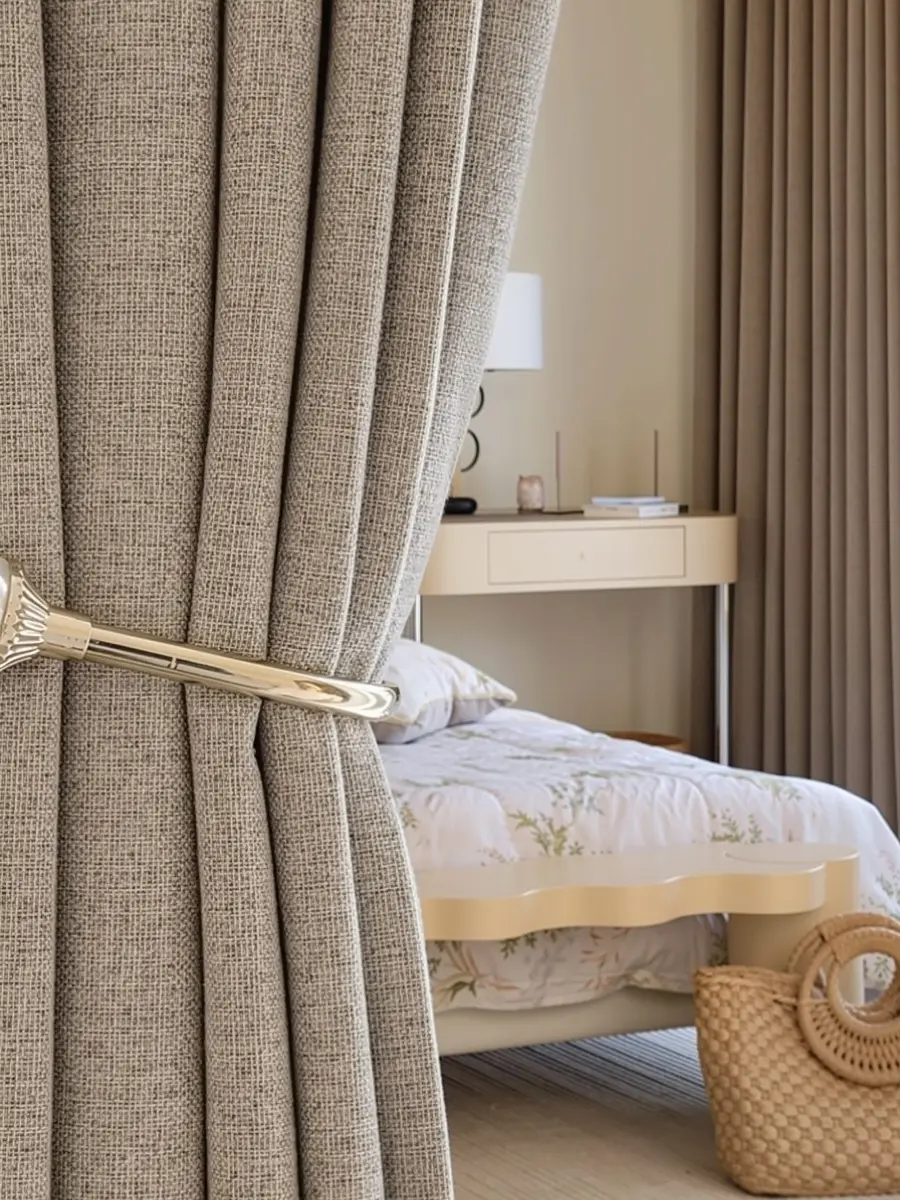 a close up of a thick, beige colored blackout curtain with a coarse, linen like texture, held by a sleek gold tieback