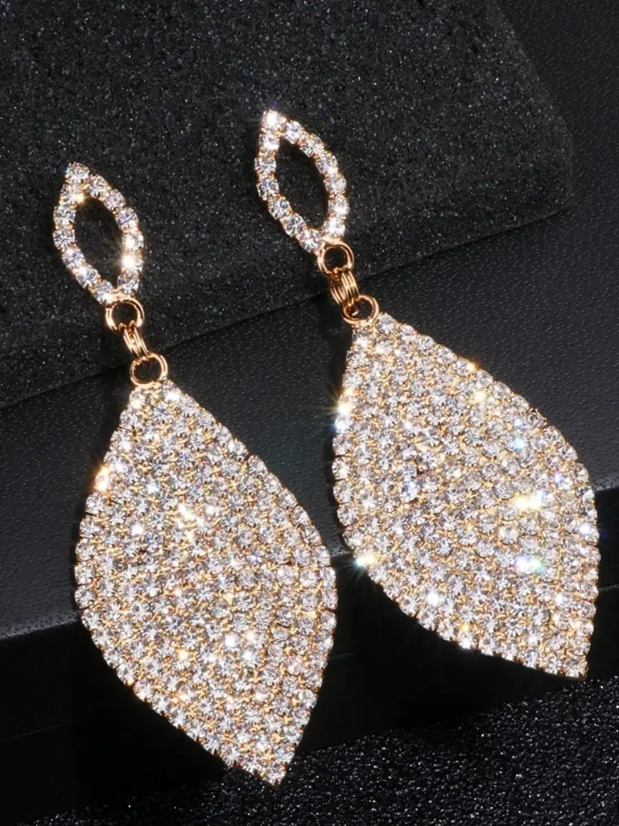 A pair of elegant, gold-toned dangle earrings encrusted with shimmering clear rhinestones. Each earring features a smaller, hollow marquise-shaped stud connected to a larger, solid leaf-shaped drop fully covered in brilliant, sparkling crystals. The earrings are displayed against a dark, textured background, highlighting their radiant glow and sophisticated design.
