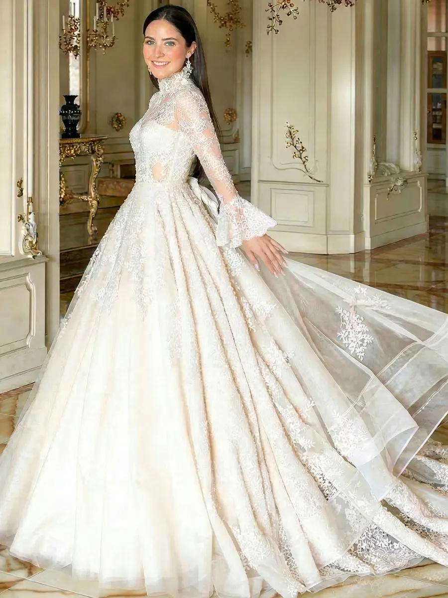A stunning, full-length bridal portrait of a woman wearing a regal, ivory-colored lace wedding gown. The dress features a modest, high-neck bodice and long sleeves that end in elegant fluted cuffs. Delicate floral lace appliques cover the top and cascade down the voluminous, multi-layered A-line skirt, which flows into a long, dramatic cathedral train. The bride is smiling as she looks over her shoulder, standing in a grand, classically designed room with ornate gold accents and tall doorways.