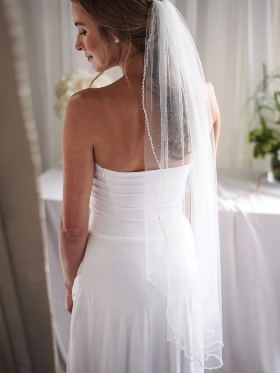 Rear view of a bride in a strapless white wedding gown wearing a delicate, sheer tulle cathedral-length veil with a shimmering beaded or rhinestone-encrusted edge.