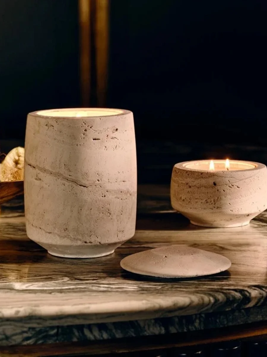 Natural travertine stone candle holders with lit candles and a matching stone lid on a marble table.