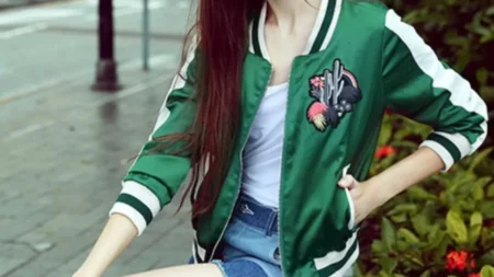 A woman wearing a green satin bomber jacket with white striped ribbed cuffs and collar. The jacket features a large colorful embroidered patch on the chest with a cactus and floral design. She is also wearing a white top and denim shorts, posing outdoors.