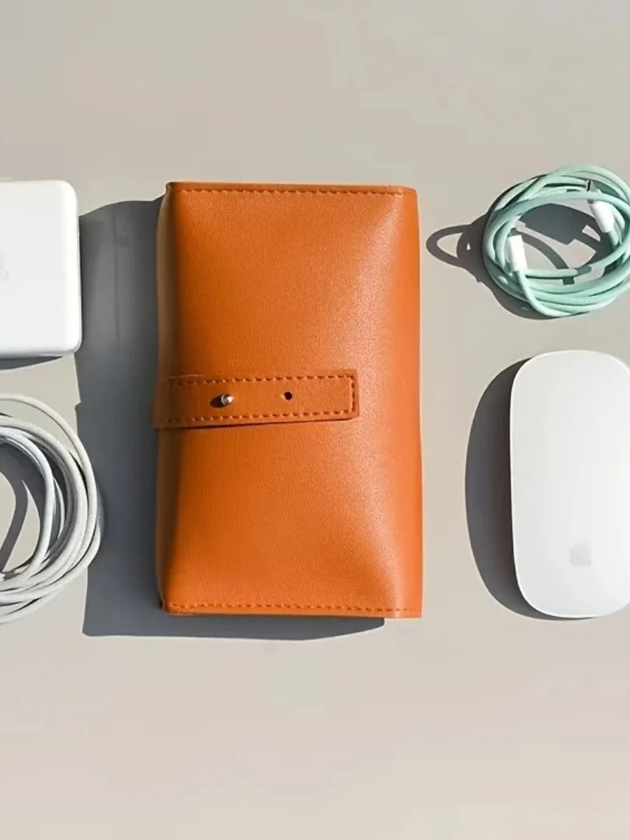 Minimalist orange PU leather tech organizer featuring a professional strap-and-stud closure, surrounded by essential travel gadgets including a white power adapter, Apple Magic Mouse, and coiled charging cables on a neutral-toned surface.