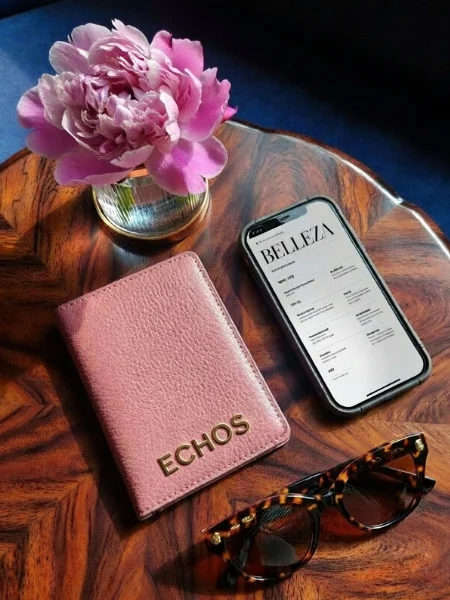 Flat lay of a pink textured leather passport holder with gold 'ECHOS' lettering, tortoise-shell sunglasses, and a blooming pink peony on a polished wood grain table, illustrating the 2026 quiet luxury lifestyle aesthetic.