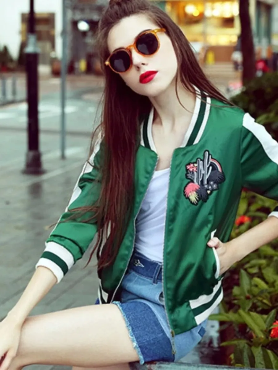 3 Affordable Luxury Spring Capsule 2026 Essentials for a 3-3-3 Wardrobe Woman wearing a green satin bomber jacket with white stripe detailing and floral embroidery, styled with denim shorts and round sunglasses for a chic, casual Spring 2026 outfit.