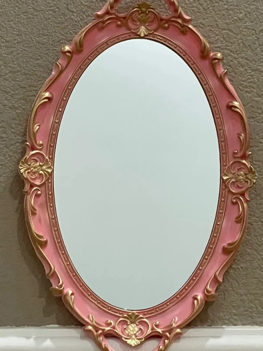 An oval, vintage-style wall mirror featuring a dusty pink frame with ornate gold rococo carvings and floral motifs. The mirror is mounted on a textured beige wall, showcasing a clean reflection.
