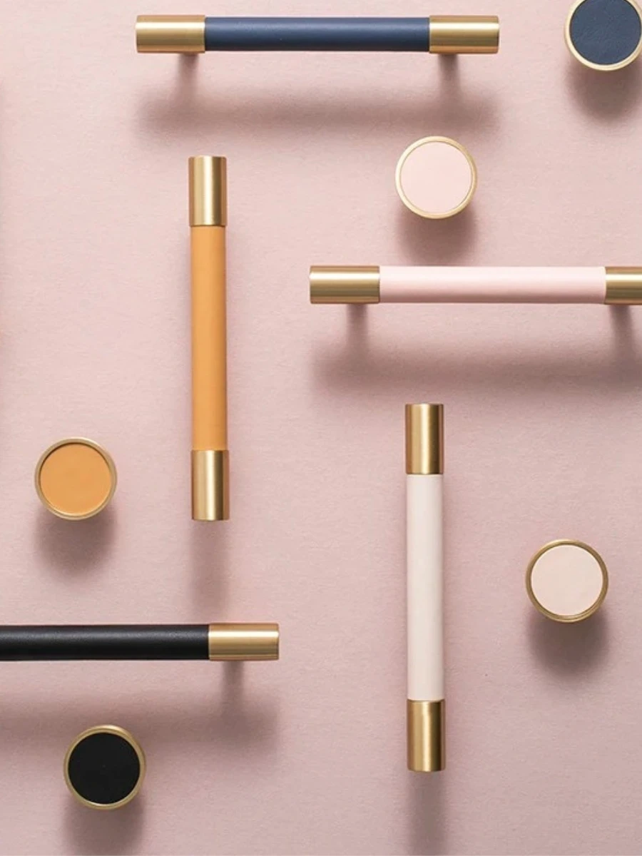 A top-down view of various modern cabinet handles and round knobs arranged artistically on a dusty pink background. The hardware features a dual-tone design with brushed gold brass ends and colorful center grips in navy blue, tan, soft pink, black, and cream.
