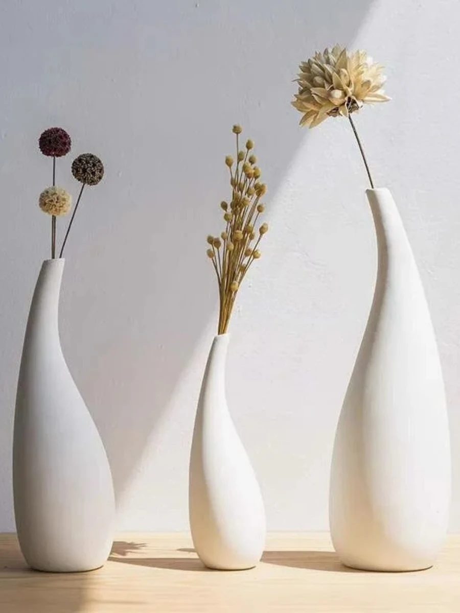 Three minimalist, teardrop-shaped white ceramic vases of varying heights and curves, each holding a single dried floral stem. They are arranged on a light wood surface against a white wall with soft, natural sunlight creating gentle shadows.