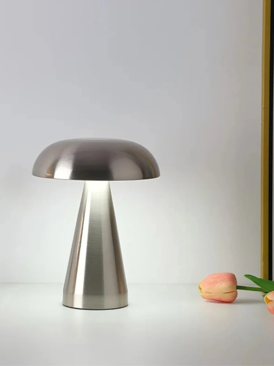 A modern silver mushroom-style table lamp with a conical base and domed shade, glowing with a soft white light on a white surface.
