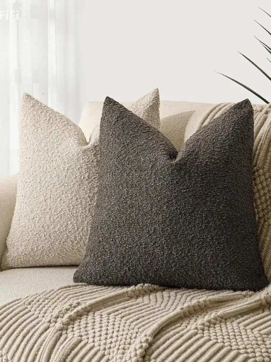 Two textured boucle throw pillows, one in cream and one in charcoal grey, arranged on a neutral sofa next to a knitted beige throw blanket.