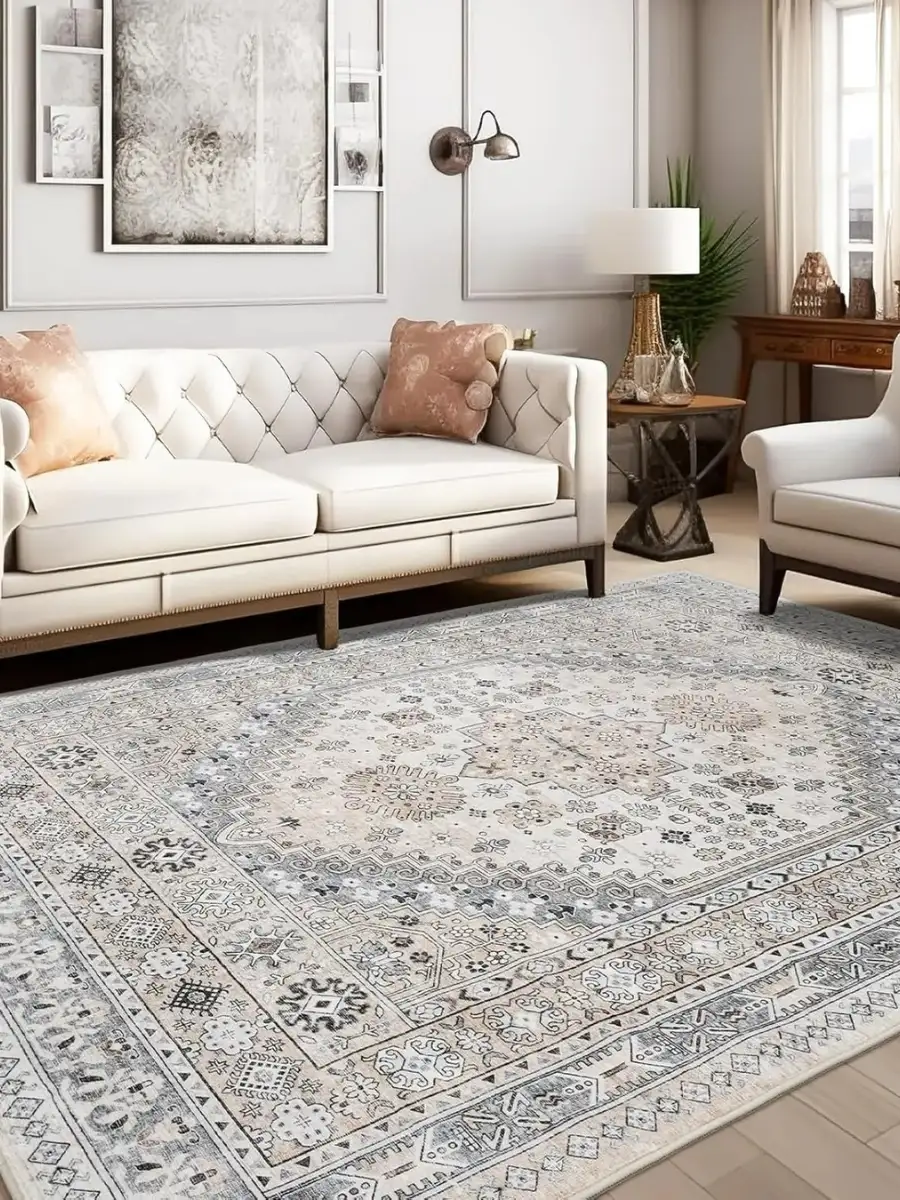 A living room setup featuring a large, light-grey distressed Persian-style area rug with intricate floral and geometric patterns. The room includes a cream-colored tufted sofa with peach pillows, a white armchair, a wooden side table with a lamp, and minimalist wall art under a brass sconce.