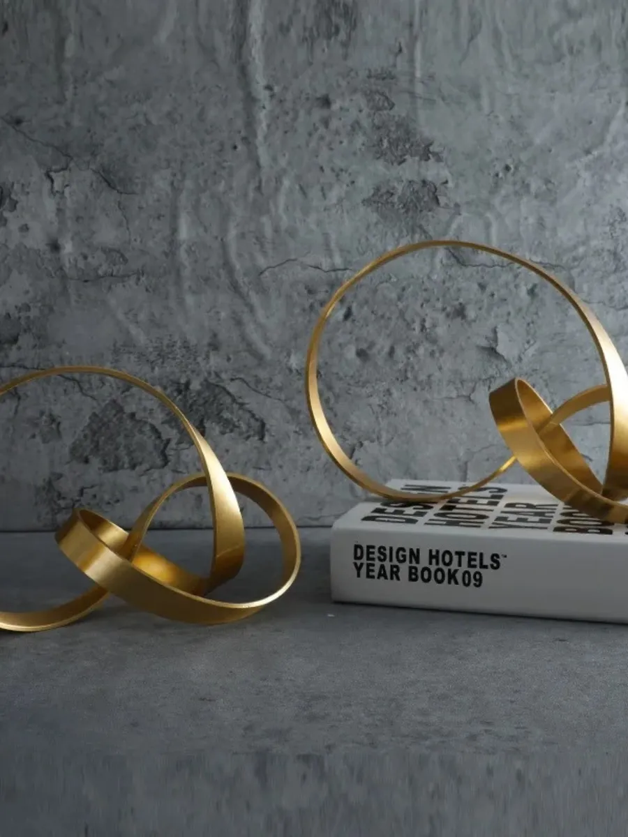 Two abstract, matte gold ribbon sculptures with interlocking loops displayed on a grey concrete-textured surface. One sculpture rests directly on the surface, while the other is placed on a white "Design Hotels Year Book 09" against a matching grey wall.