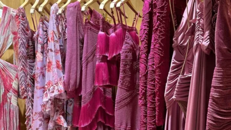 A row of elegant blush, soft rose, and mauve formal midi dresses hanging on wooden garment hangers inside a luxury boutique wardrobe closet.