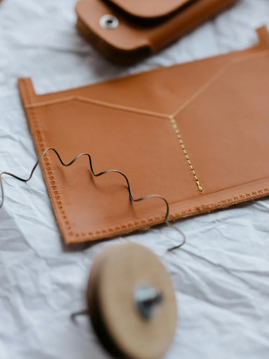 A close-up view of handmade leather goods in a workshop setting, featuring a stitched tan leather pouch envelope sleeve with yellow thread work resting on wrinkled white paper alongside craft tools.