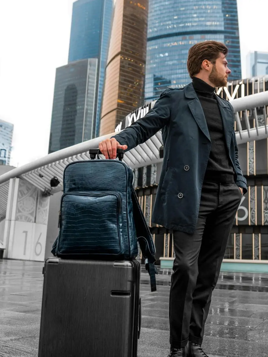 A man in a navy blue trench coat and black turtleneck standing in a modern city plaza with skyscrapers, resting his hand on a blue crocodile-embossed leather backpack that is stacked on top of a black hardshell rolling suitcase.