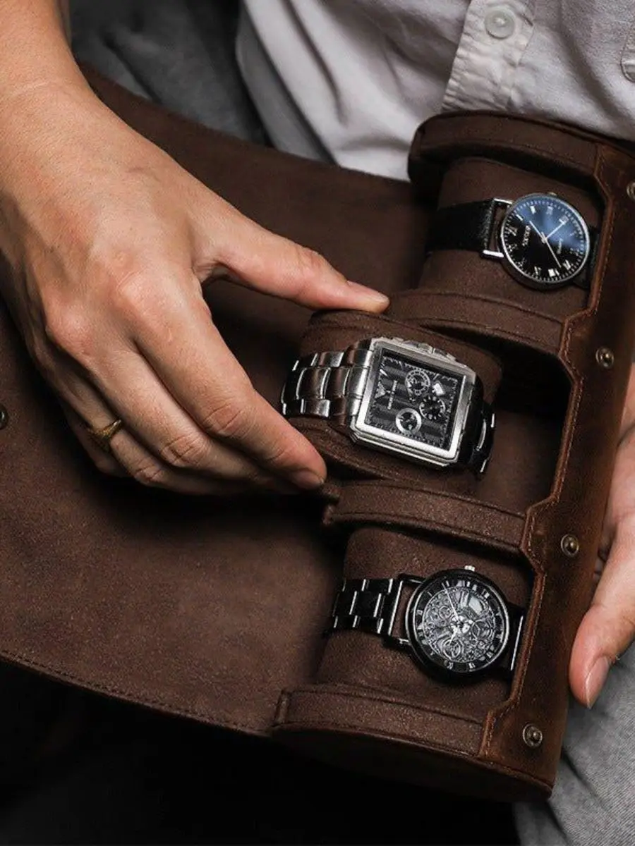 A person’s hands organizing three luxury wristwatches inside a brown leather travel watch roll case with soft suede compartments.