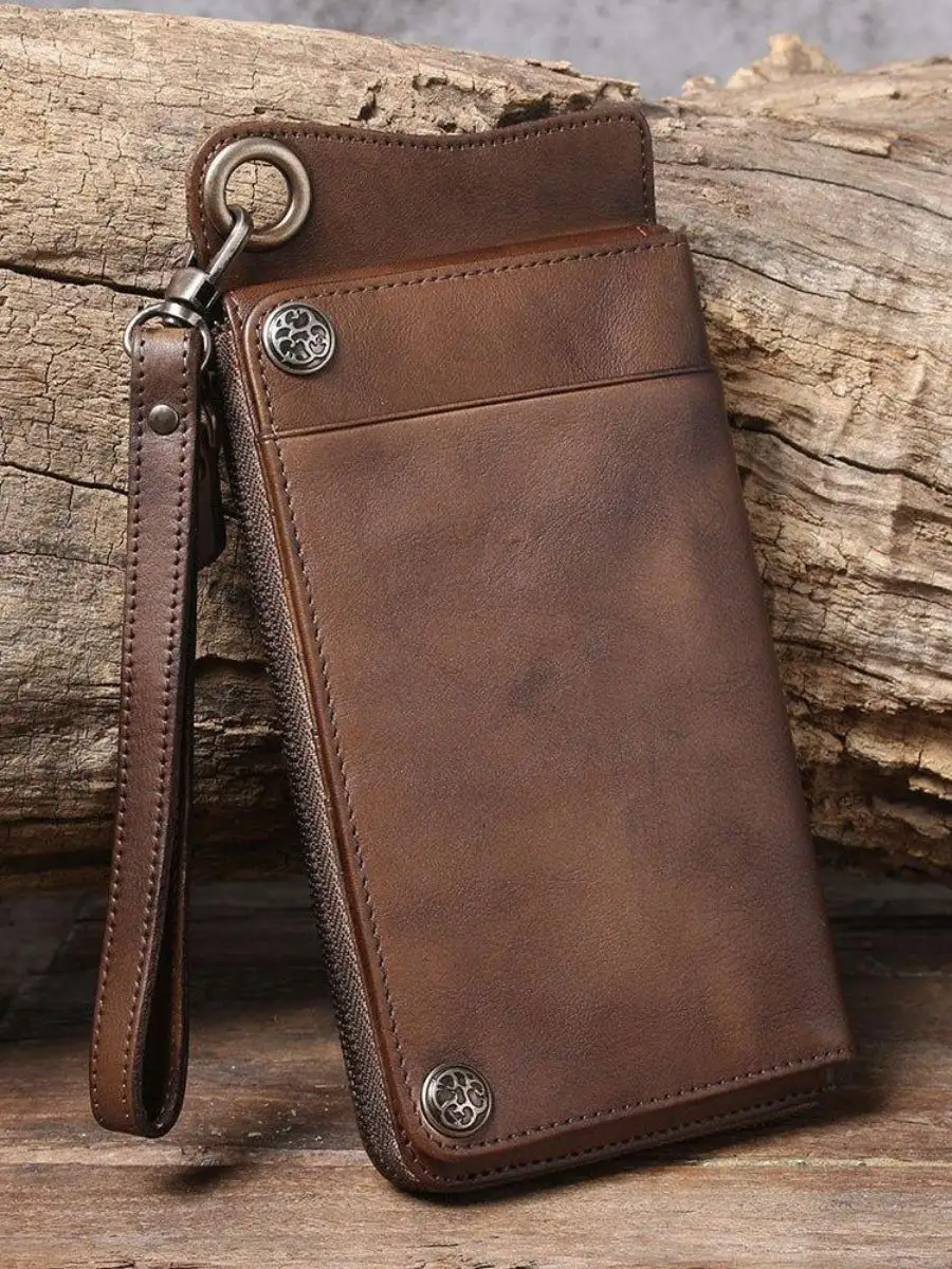 A long vintage brown leather men's wallet featuring silver metallic snap fasteners, a side zipper closure, and a matching leather wrist strap against a rustic wood background.