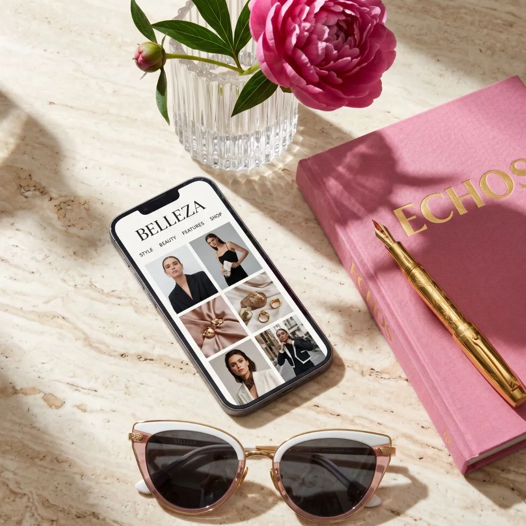 Luxury editorial flat lay featuring a smartphone displaying the Belleza Echos lifestyle blog alongside a premium pink hardcover notebook, a solid gold fountain pen, a fresh pink peony, and elegant white and rose gold cat-eye designer sunglasses on a travertine marble surface.