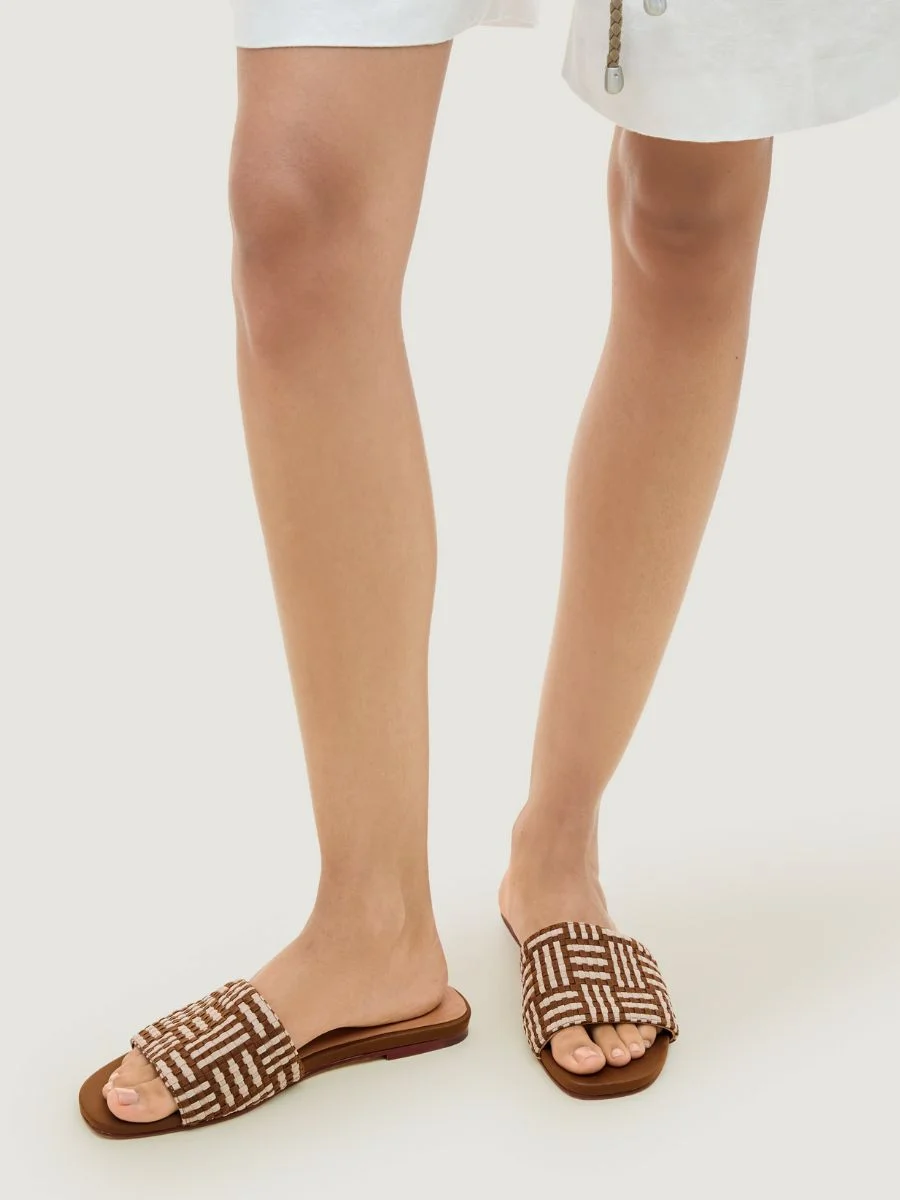 A close-up view of a person wearing the Cartolina Nantucket Cairo Slide in Almond, featuring a brown and cream geometric woven strap and a flat leather footbed.