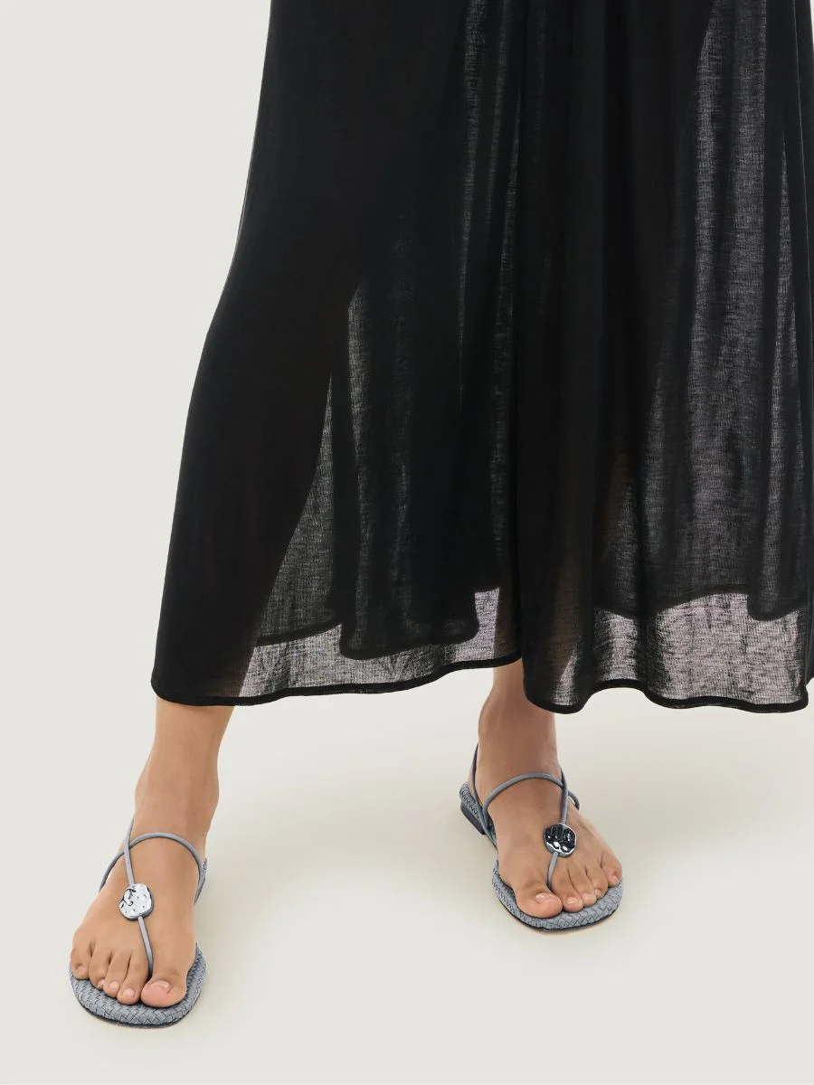 A woman wearing a long black linen skirt and the Cartolina Nantucket Gaia Sandal in Marina blue, featuring thin cord straps and a hammered metallic disc.