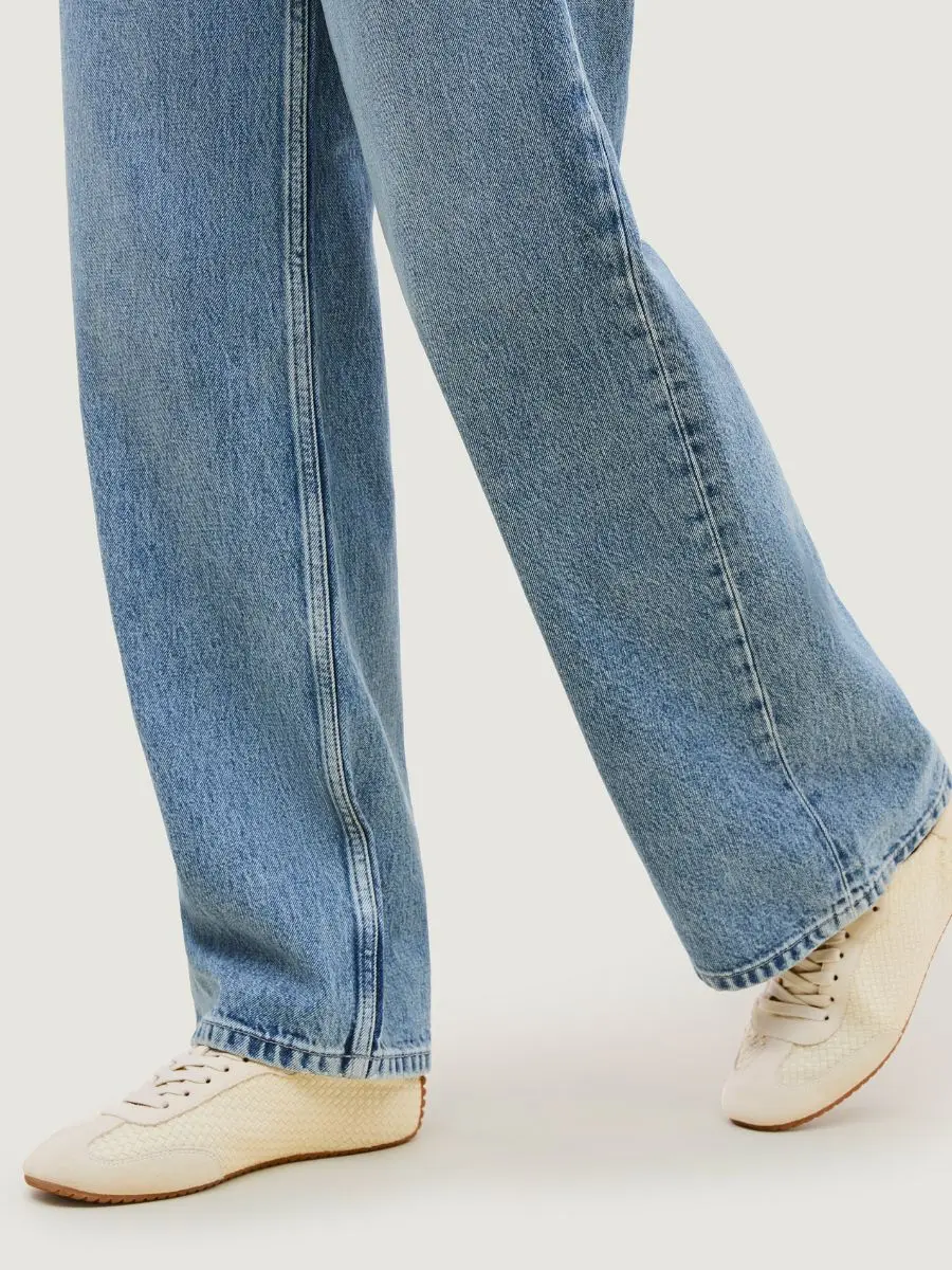 A close-up view of a person wearing wide-leg blue jeans and the Cartolina Nantucket Milan Sneaker in bone white handwoven neoprene and suede.