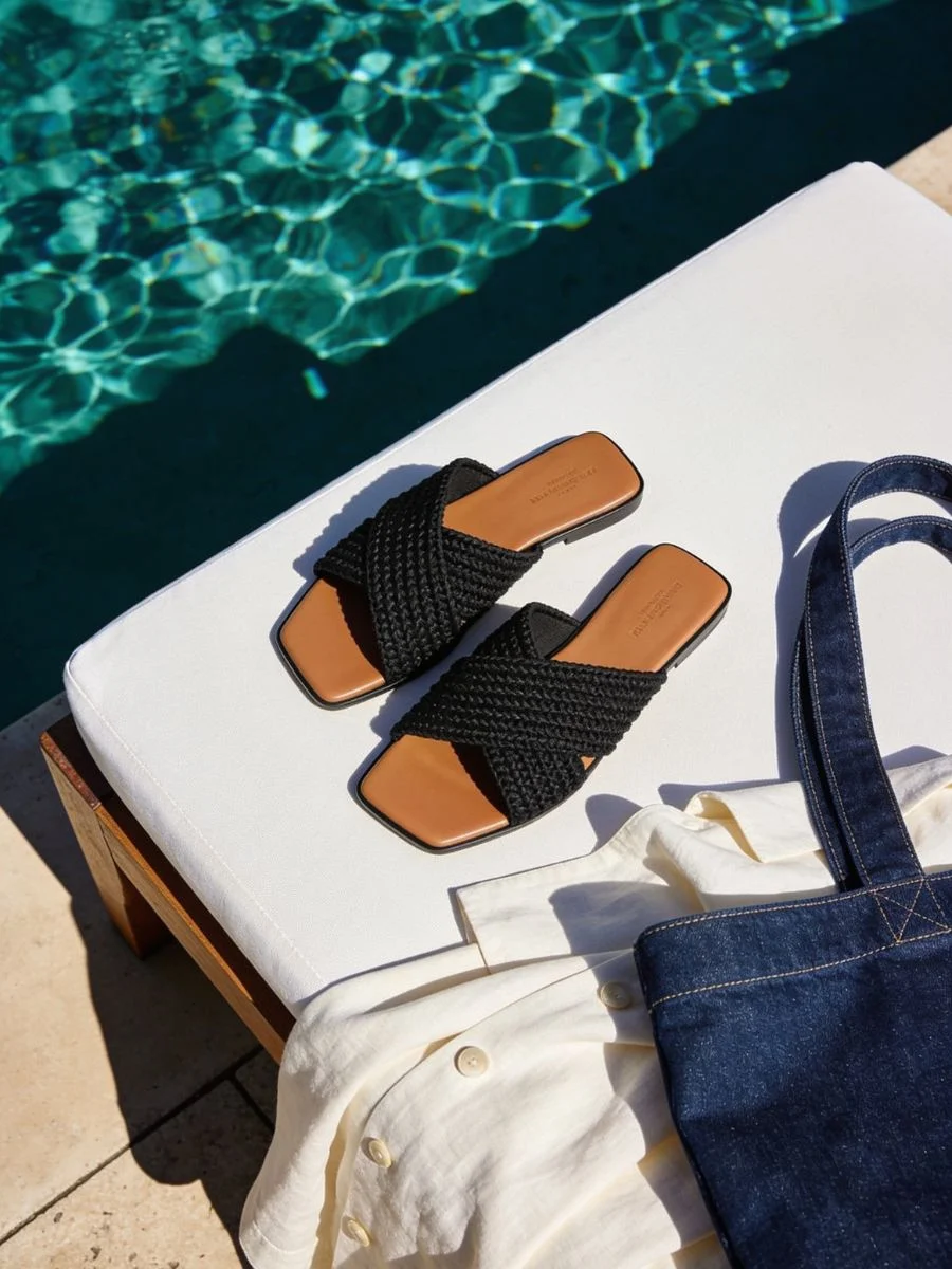 A pair of elegant black woven neoprene summer resort slide sandals resting on a sun lounger next to deep turquoise pool water, styled with a denim tote and linen shirt.