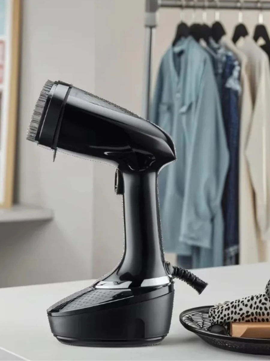 A sleek, minimalist black handheld clothes steamer resting on a clean white vanity table, positioned in front of an elegant open capsule wardrobe with curated luxury garments hanging on sleek black hangers.