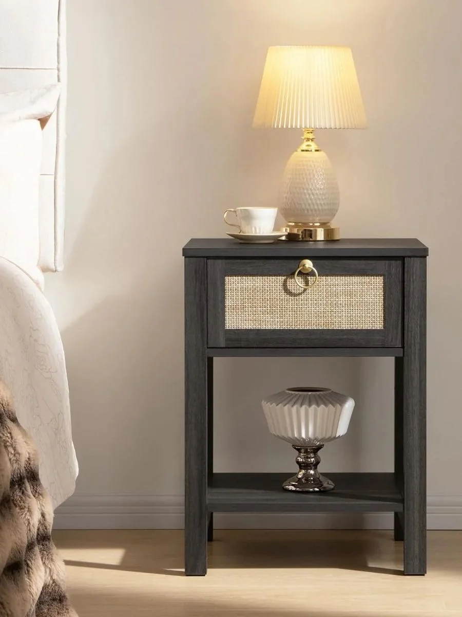A dark charcoal bedside table featuring a natural cane drawer front and a polished gold ring pull handle, styled with a glowing table lamp.