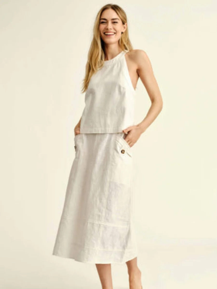 A woman stands smiling on a white background wearing an oatmeal linen halter-style sleeveless separate top and a matching high-waisted linen midi skirt with side button flaps.