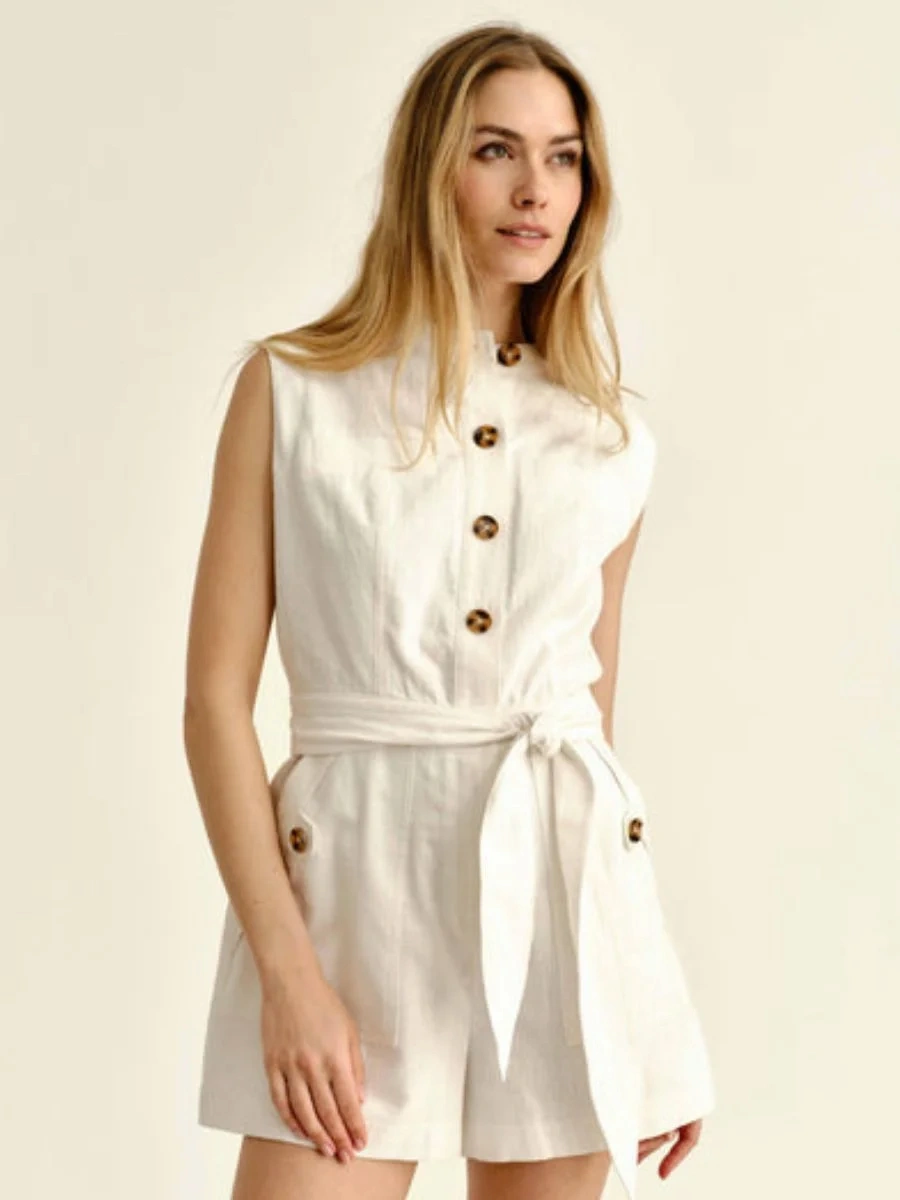A woman wearing a sleeveless white cotton-blend resort romper with brown buttons for a classy white outfit on a tropical holiday.
