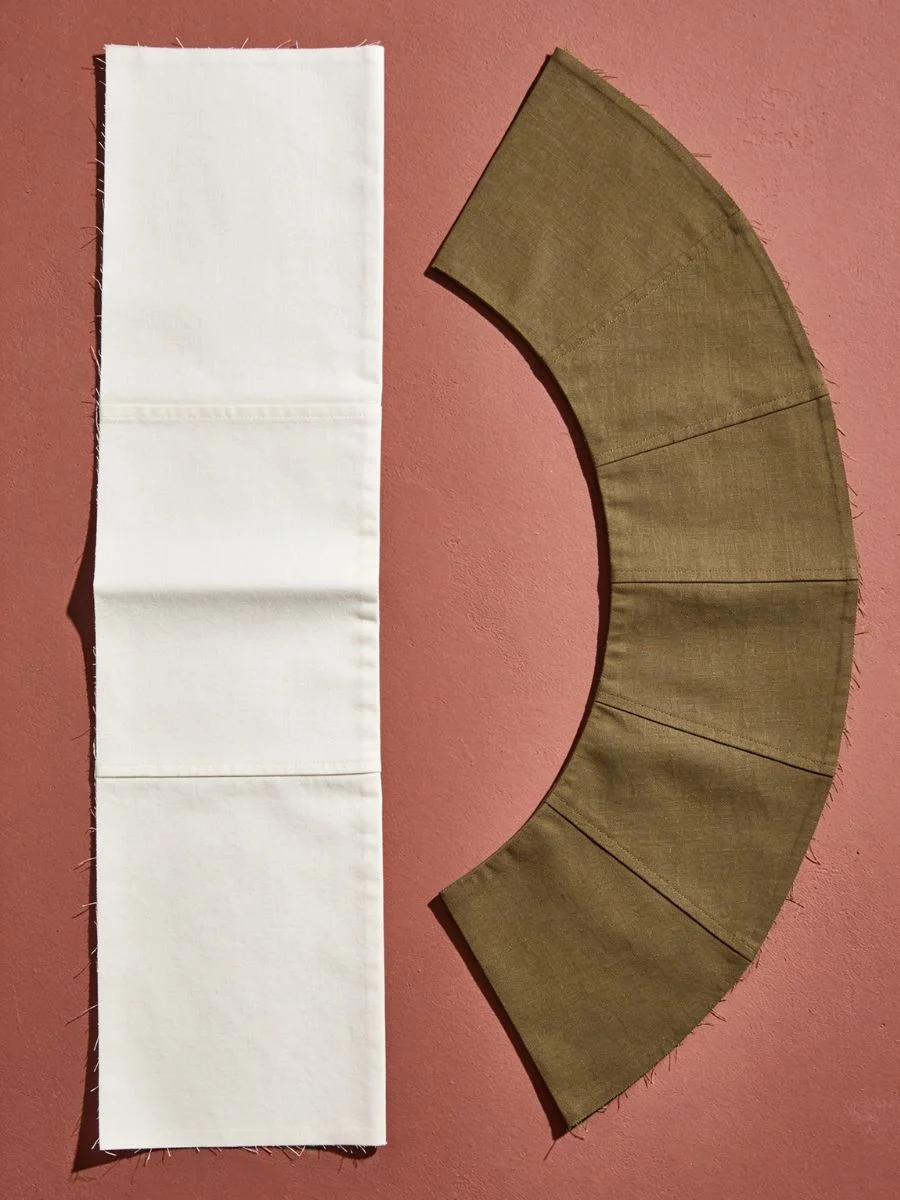 Flat white rectangular canvas fabric strip next to a curved dark khaki crescent arc waistband template on a terracotta surface.