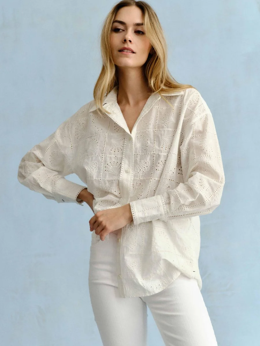 A woman stands against a light blue backdrop wearing a soft white Cartolina Margaux long-sleeved button-down shirt featuring an all-over geometric eyelet design, paired with fitted white pants.