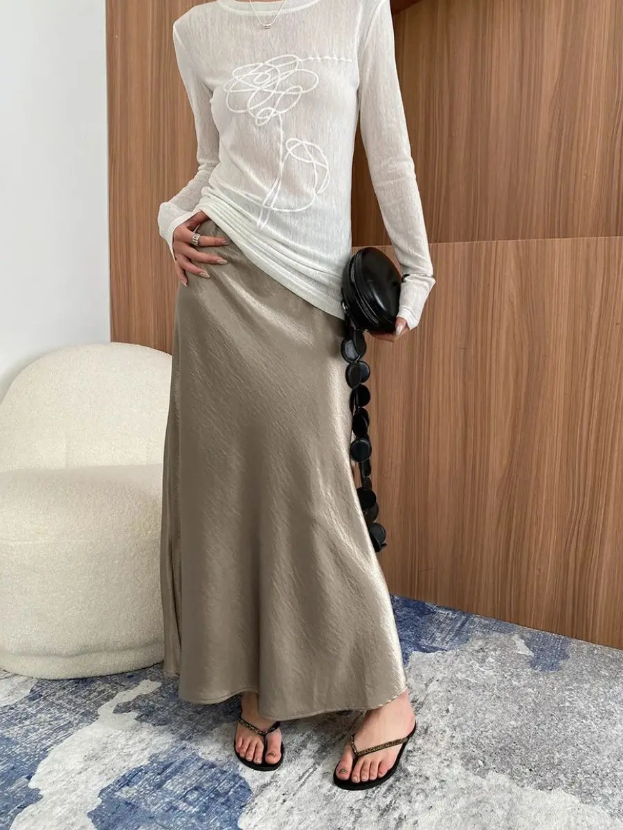 An elegant monochromatic outfit featuring a model wearing a champagne satin midi skirt paired with a long-sleeve semi-sheer white top, holding a modern black structural bag