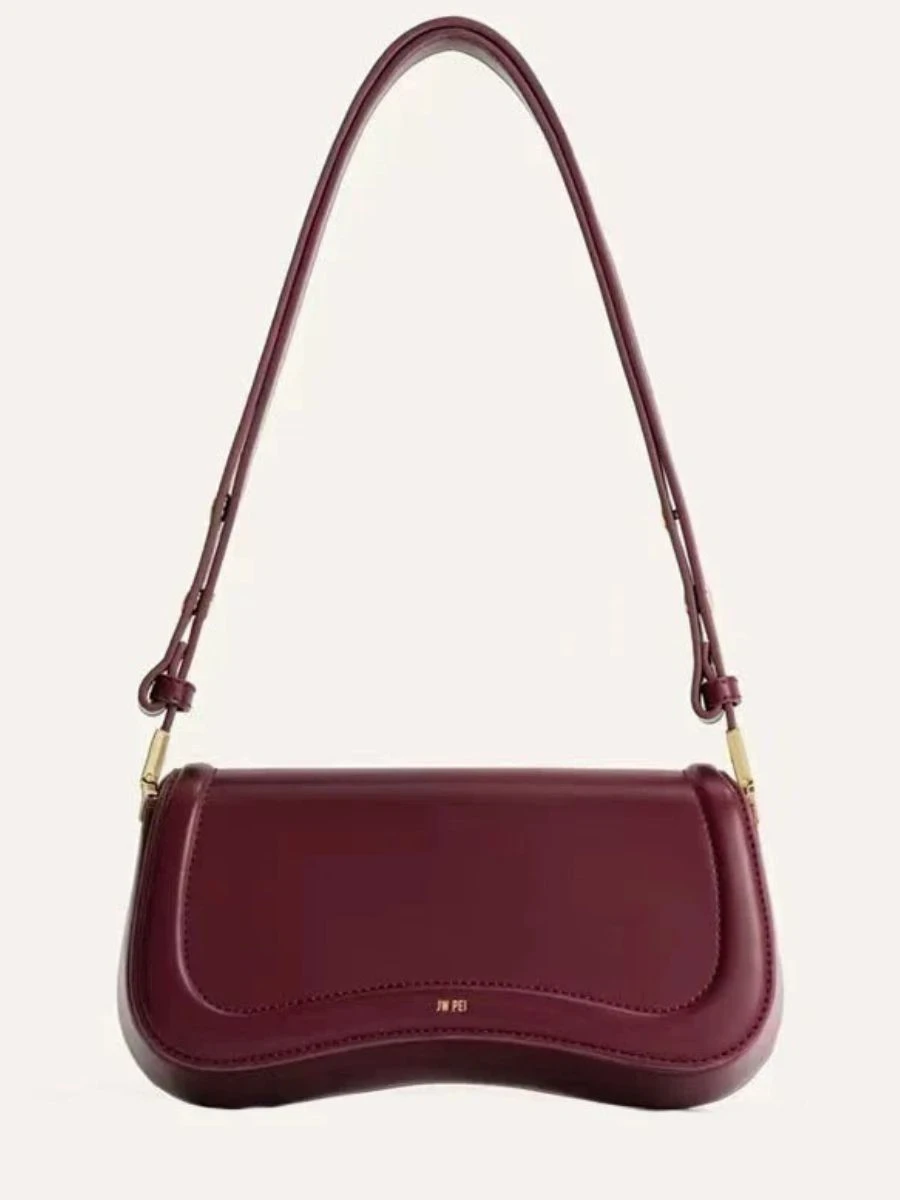 A structured burgundy vegan leather shoulder bag with a sleek architectural curved base and a minimalist gold stamp accent against a plain white backdrop.