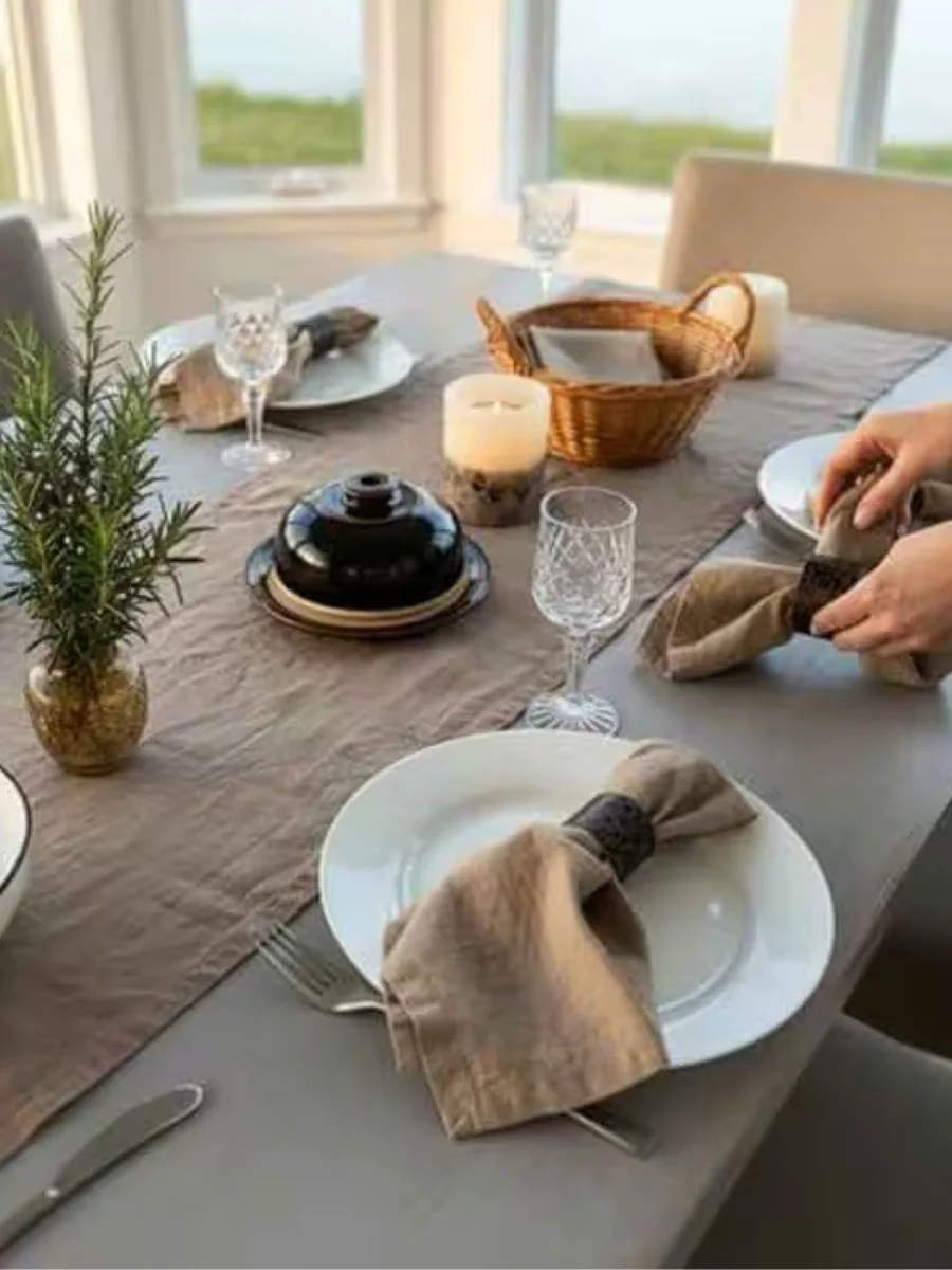 Hands setting a dining table with a natural taupe 100% French flax linen table runner and matching cloth napkins in dark wooden rings.