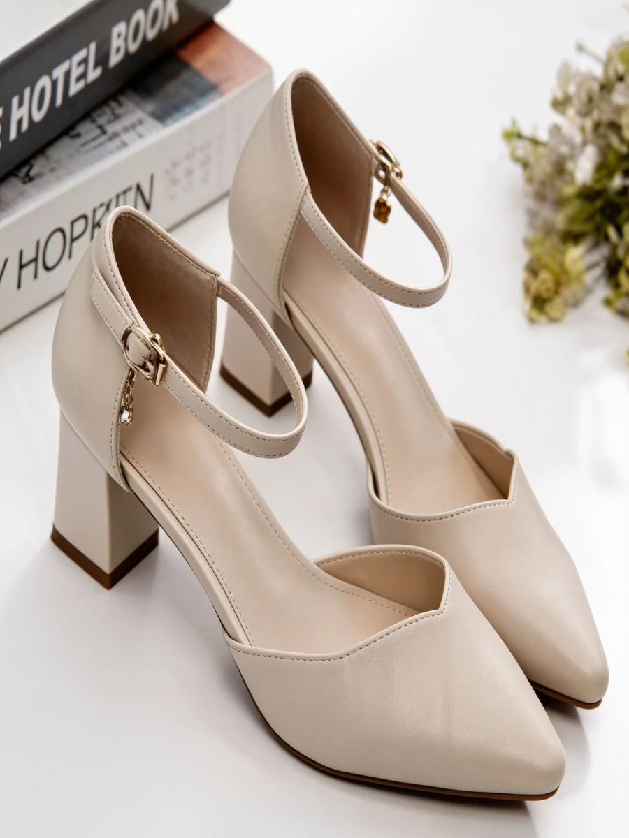 A pair of elegant cream pointed-toe d'Orsay pumps with a comfortable block heel and delicate ankle straps, styled neatly on a reflective tabletop next to decor books.