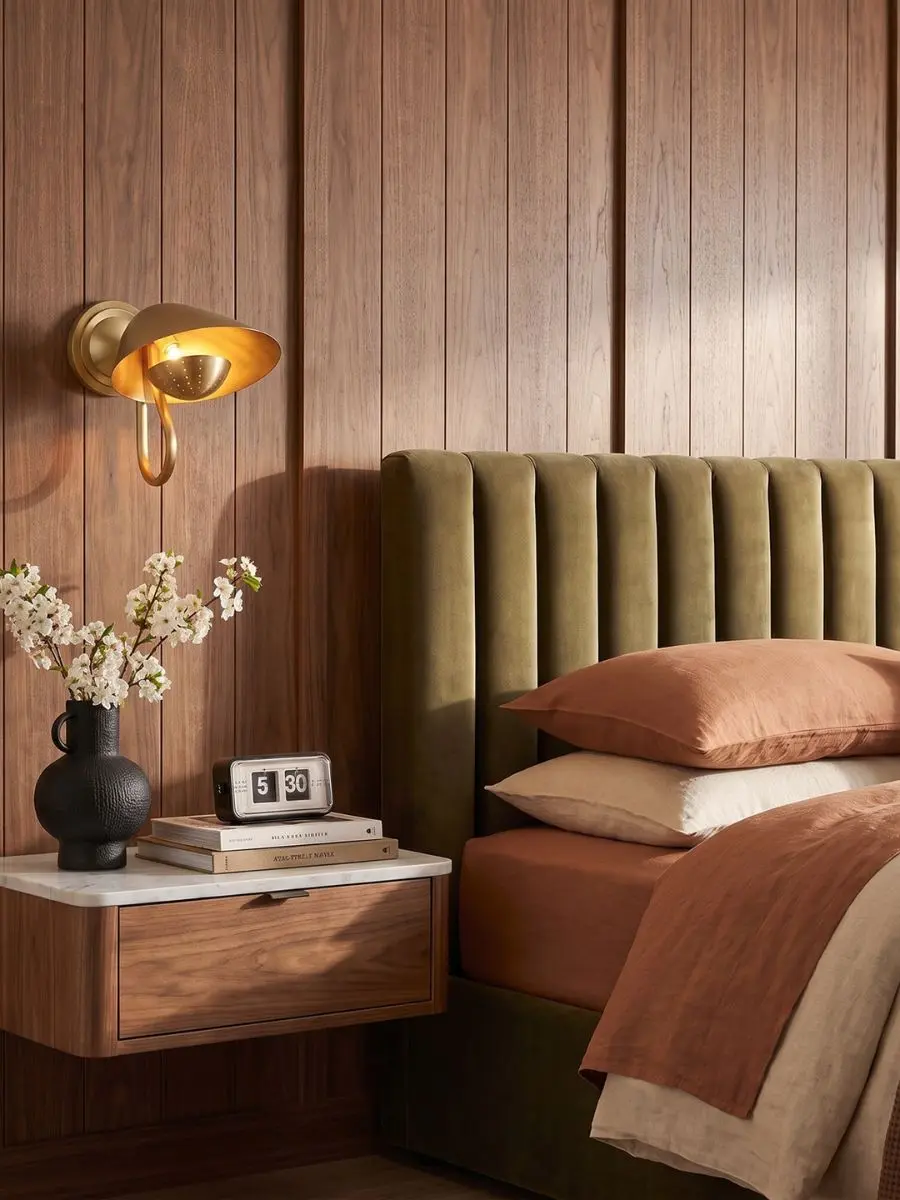 7 Layered Lighting Looks: How to Style Elegant Wall Sconces and Lamps Regina Andrew Keaton Single Sconce in natural brass mounted above a bedside table in a bedroom with wood paneling.