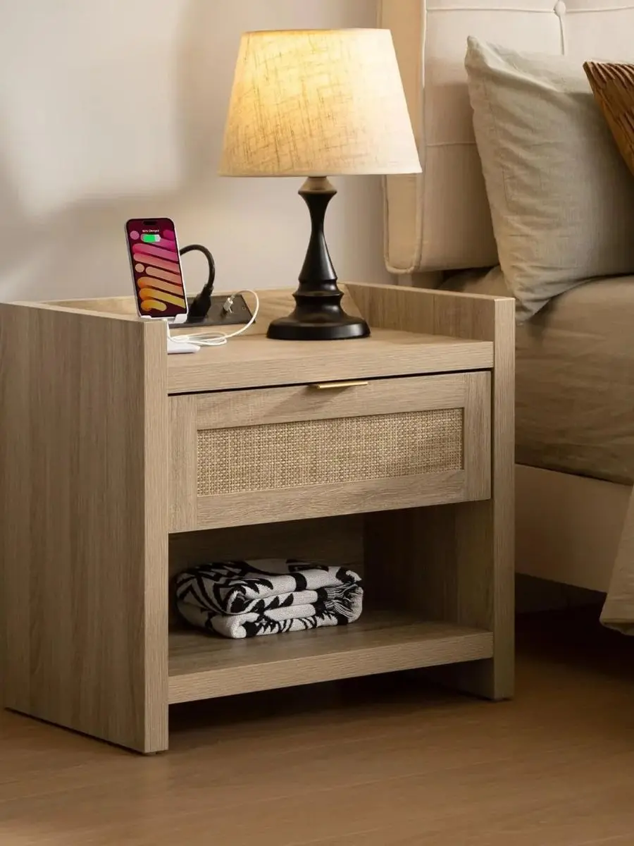A light oak cane nightstand with gold hardware featuring a built-in power strip charging a phone next to an illuminated warm bedside lamp.