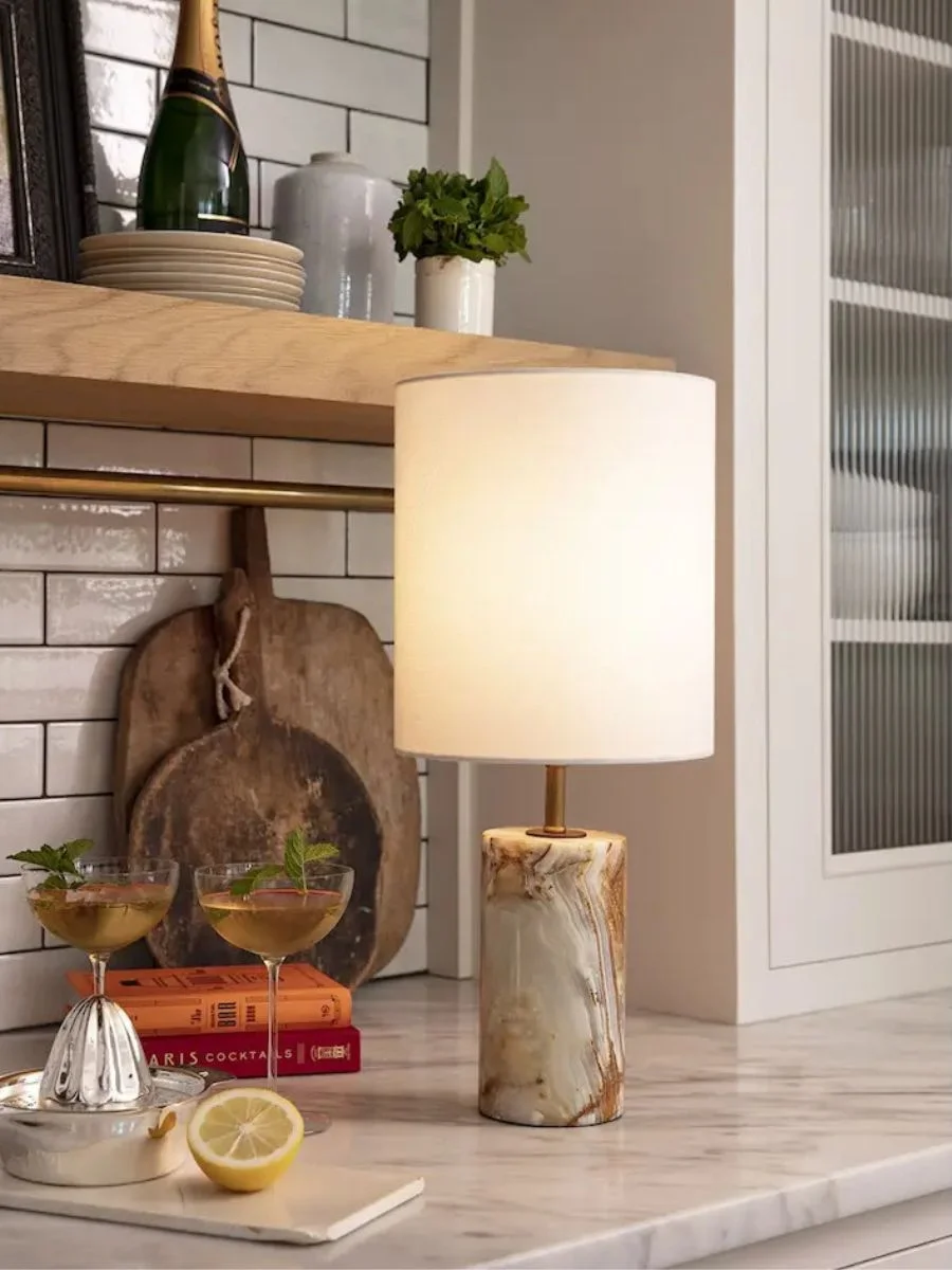 7 Layered Lighting Looks: How to Style Elegant Wall Sconces and Lamps Regina Andrew Jade and Brass Mini Cylinder Lamp with a white shade styled on a marble kitchen counter next to cocktails.