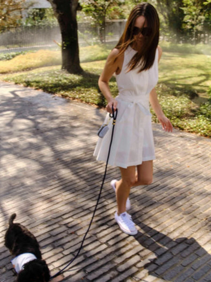 A woman walks her dog outdoors wearing a bright white, sleeveless pleated resort romper with a fabric waist tie and white sneakers.
