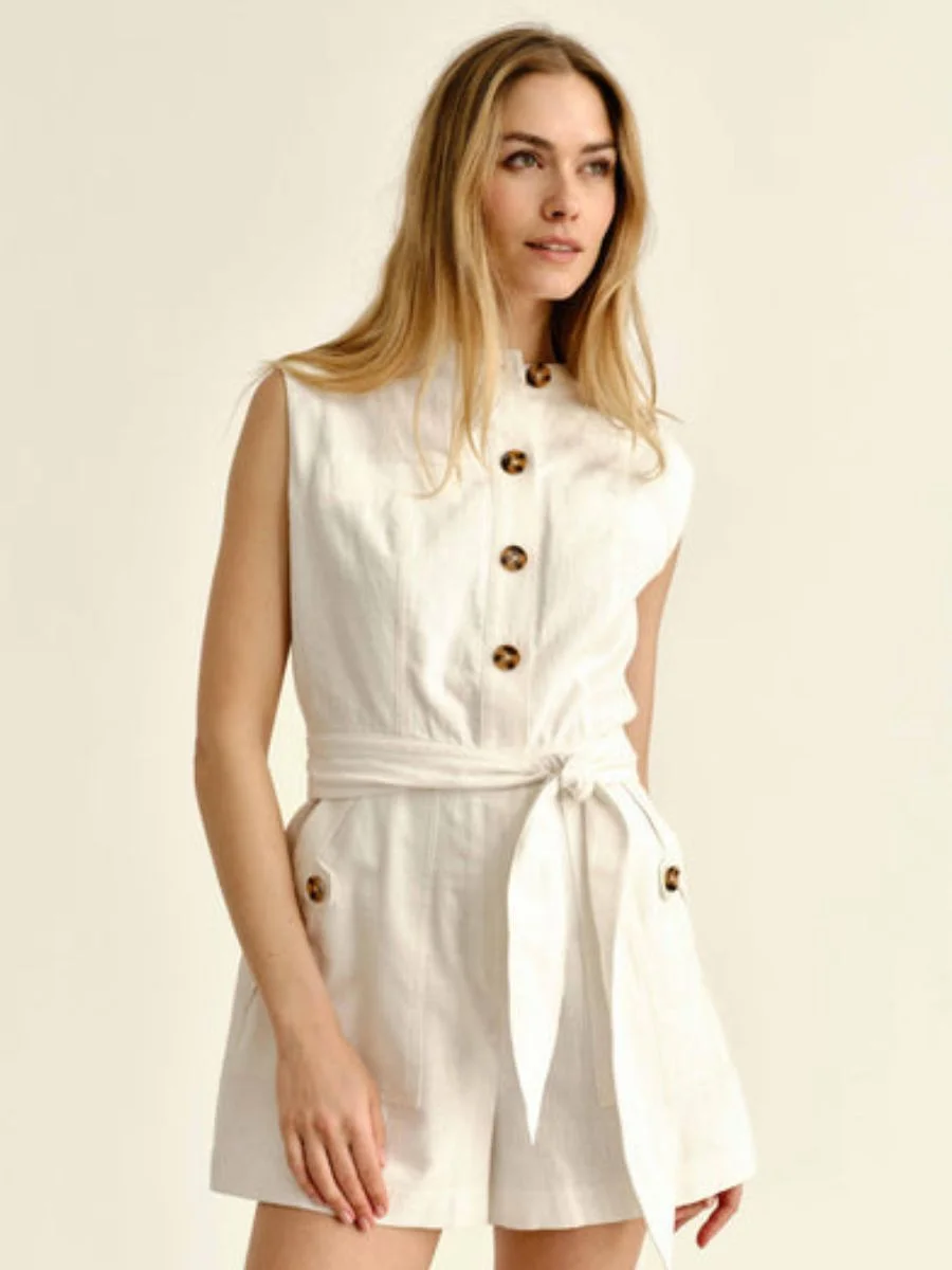  A woman stands against a cream background wearing the ivory Cartolina Rachel sleeveless mock-neck resort romper featuring a front column of brown buttons, side pocket tabs, and a matching fabric waist belt.