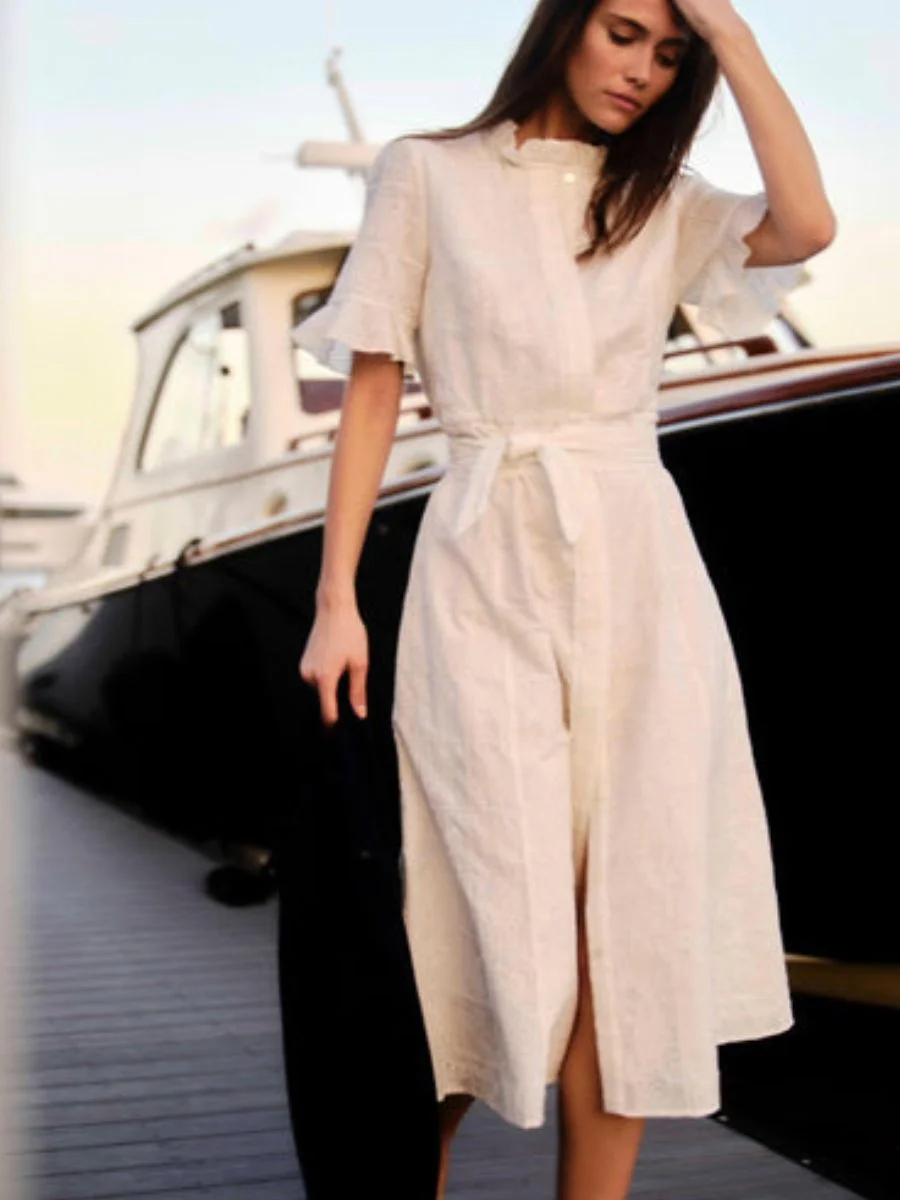  A woman stands on a wooden marina dock next to a yacht wearing the soft white Cartolina Aria midi shirt-dress featuring embroidered eyelet details, a ruffled high collar, flutter sleeves, and a fabric waist tie.