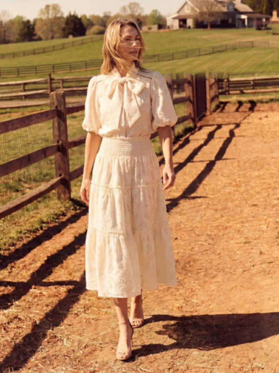 A woman walks outdoors on a sunny ranch path wearing the ivory Cartolina Peggy puff-sleeve bow neck blouse tucked into the matching tiered ivory Cartolina Kylie midi skirt.