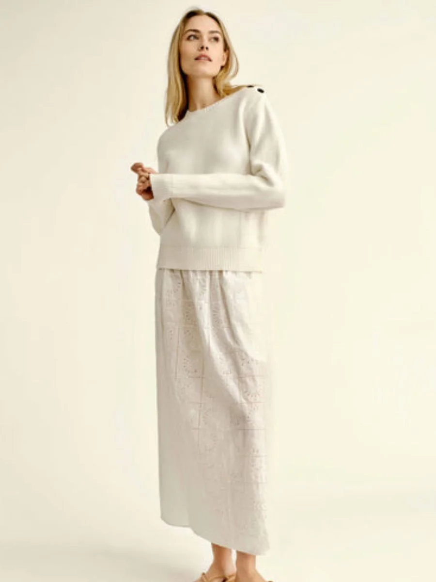 A woman stands against a light cream background wearing a soft white Cartolina Collins long-sleeved knit sweater with button details on the shoulder, styled over the matching white embroidered Cartolina Celine eyelet skirt with flat tan leather flip-flop sandals.